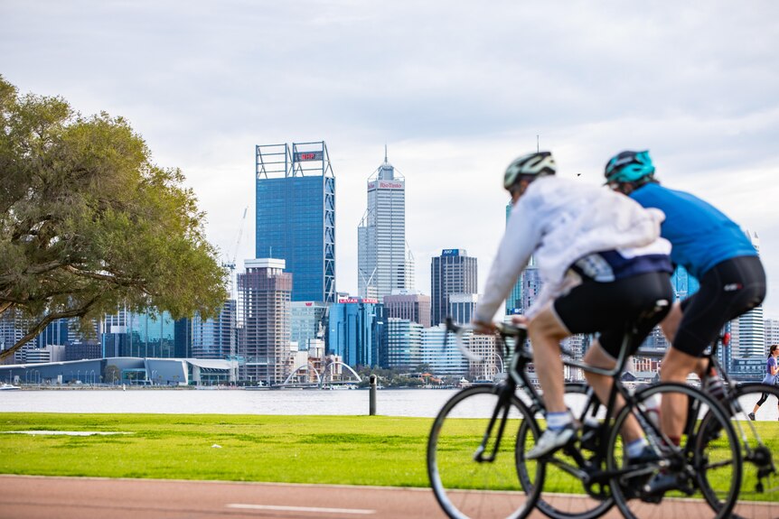 Two cyclists riding along a path on a river foreshore with a city skyline in the background.
