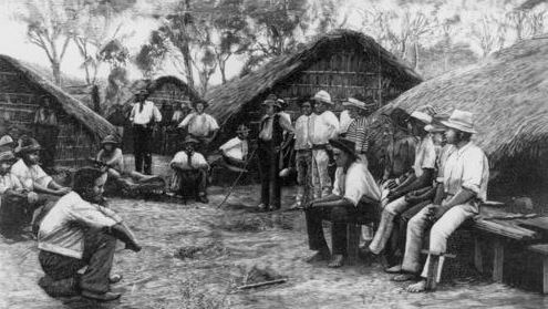 A black-and-white image shows a group of South Sea Islander labourers sitting and standing outside grass huts.