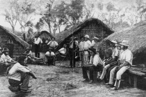 A black-and-white image shows a group of South Sea Islander labourers sitting and standing outside grass huts.