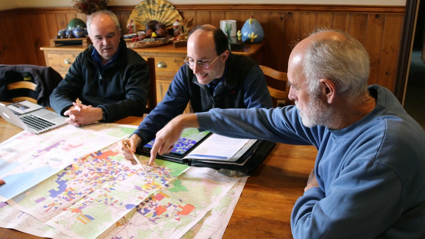 Three men sit around a table in a home and point to a map.