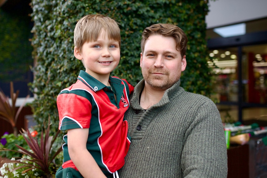 A man with some stubble holds his primary-school-aged son, wearing a school uniform.
