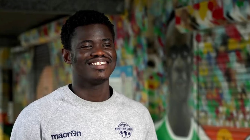A dark-skinned man wearing a grey club pullover and a wide smile. He's standing near a wall painted with lots of colours