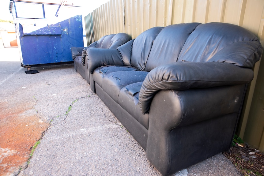 Two ripped and damaged couches sit in a bitumen yard next to a skip