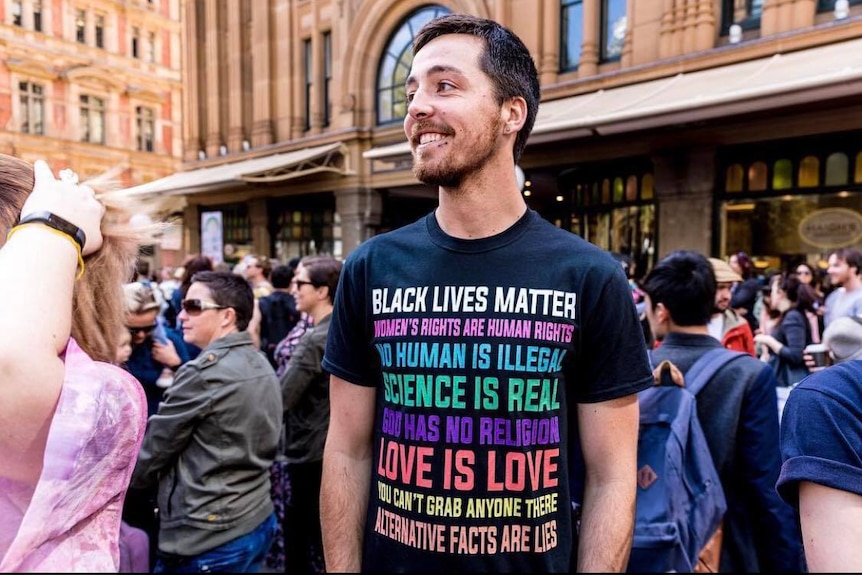 A man wearing a black graphic t-shirt stands in a crowd posing for a photo