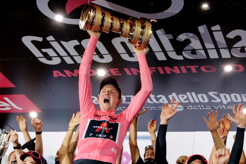 Tao Geoghegan Hart holds the trophy above his head and yells in celebration. He's wearing the pink jersey