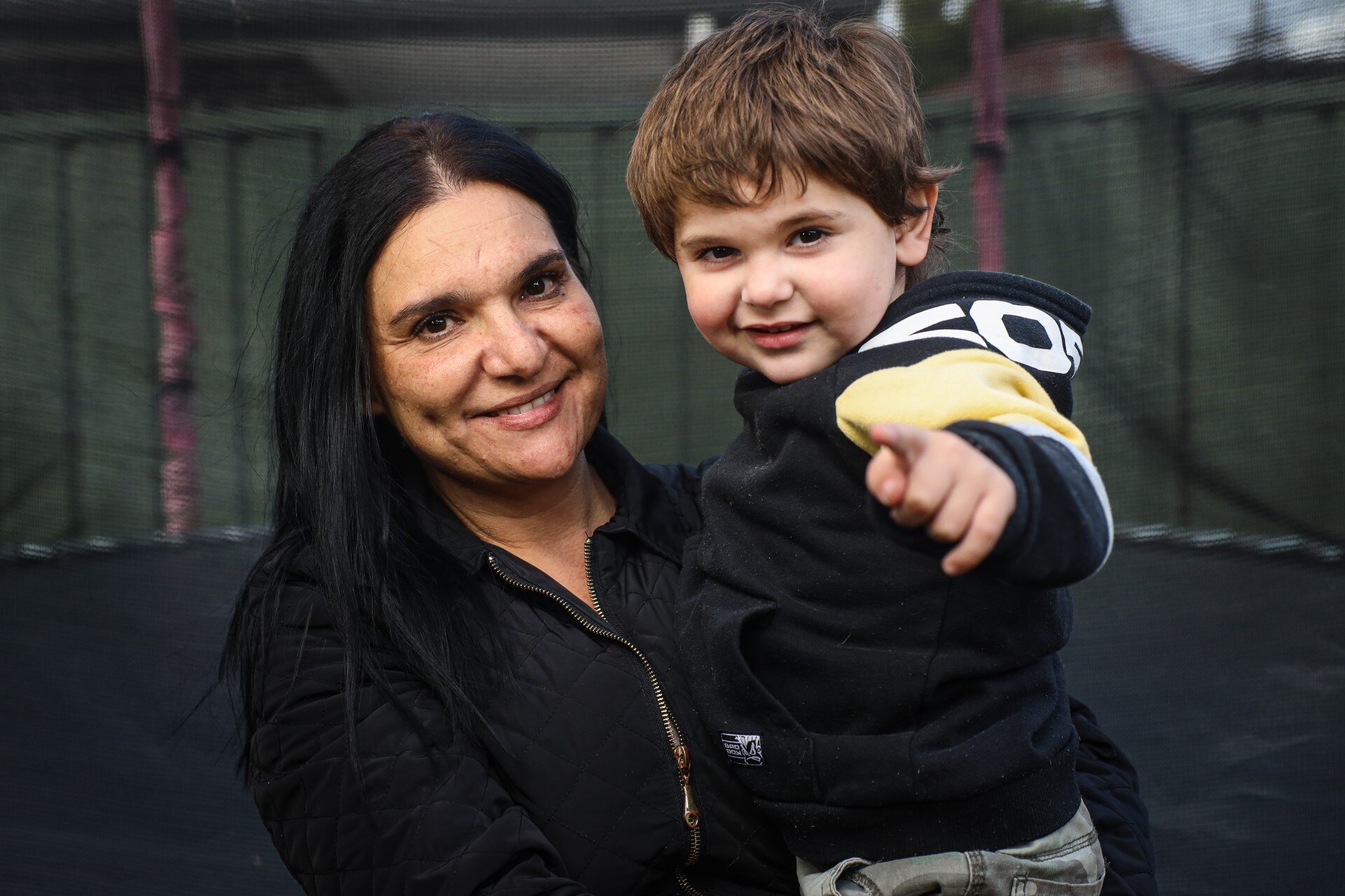 A woman with dark hair smiles as she poses for a photo with her young son in front of a trampoline.