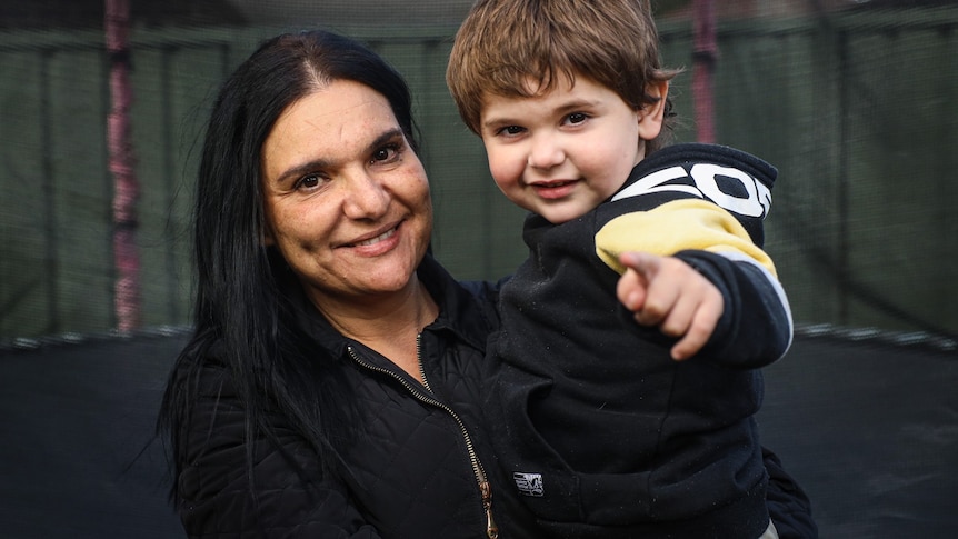 A woman with dark hair smiles as she poses for a photo with her young son in front of a trampoline.