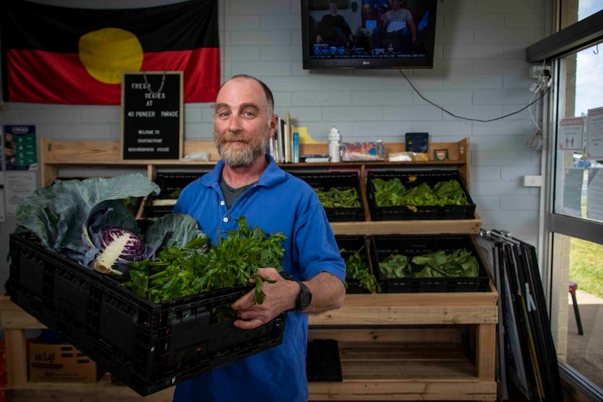 A balding man in a blue shirt holds a box of leafy vegetables