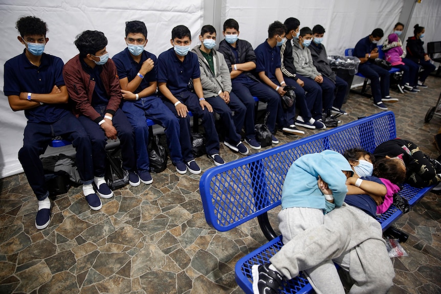 Some children sit on benches while others sleep in a crowded space