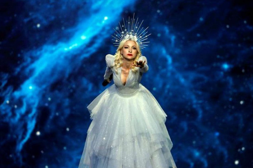 Kate Miller-Heidke is reaching towards the camera, suspended on a pole, with a microphone in opposite hand.