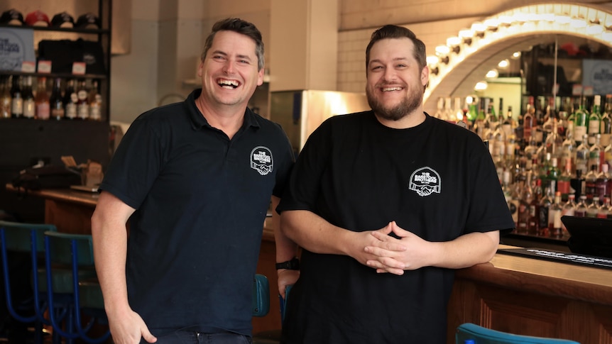 Lachlan Stevens and Daniel Chin laugh while standing in front of a bar, wearing black shirts.