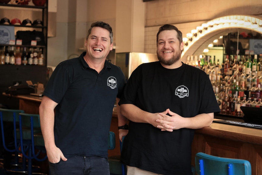 Lachlan Stevens and Daniel Chin laugh while standing in front of a bar, wearing black shirts.