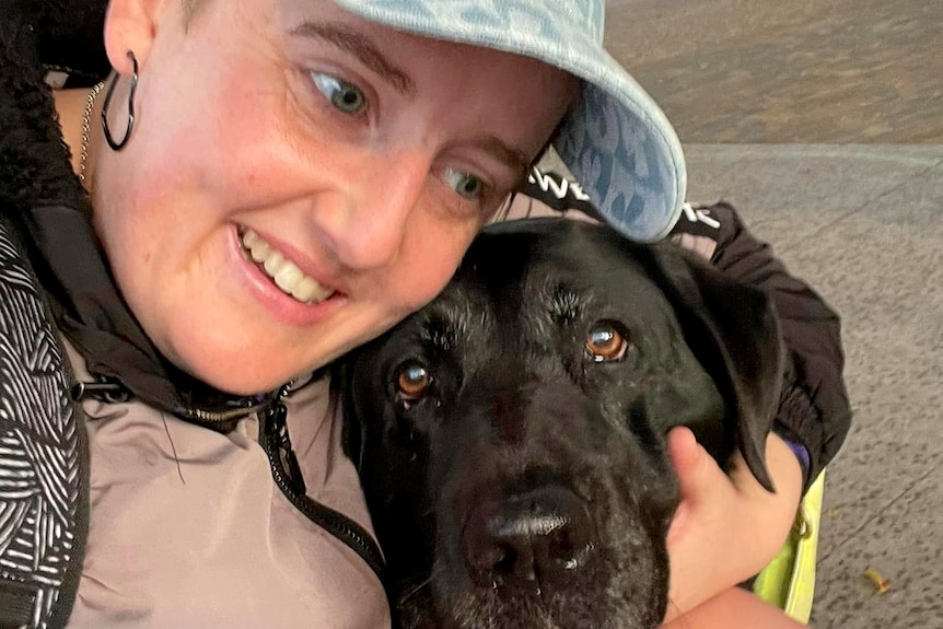 Smiling woman cuddling a black labrador that is looking at the camera