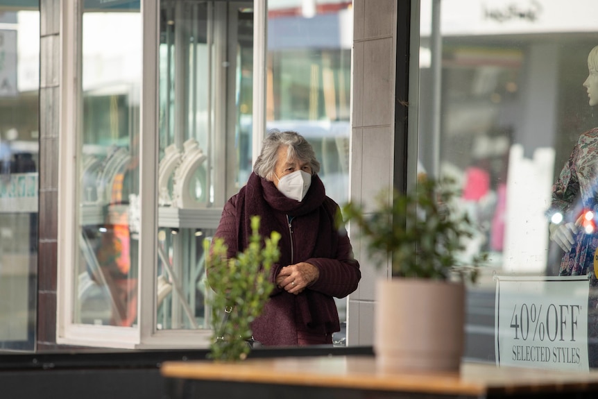 A woman walking past shops on a city street, wearing a face mask