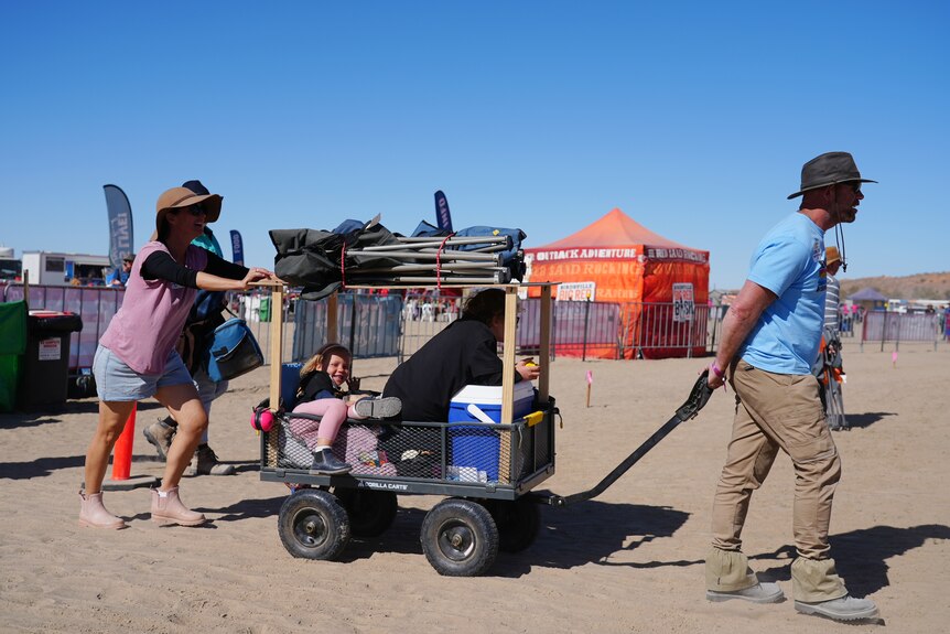 A man drags a luggage cart containing an Esky and child in the desert.