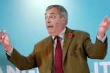 Nigel Farage wears a brown jacket, gestures with his hands in the air and a look of surprise