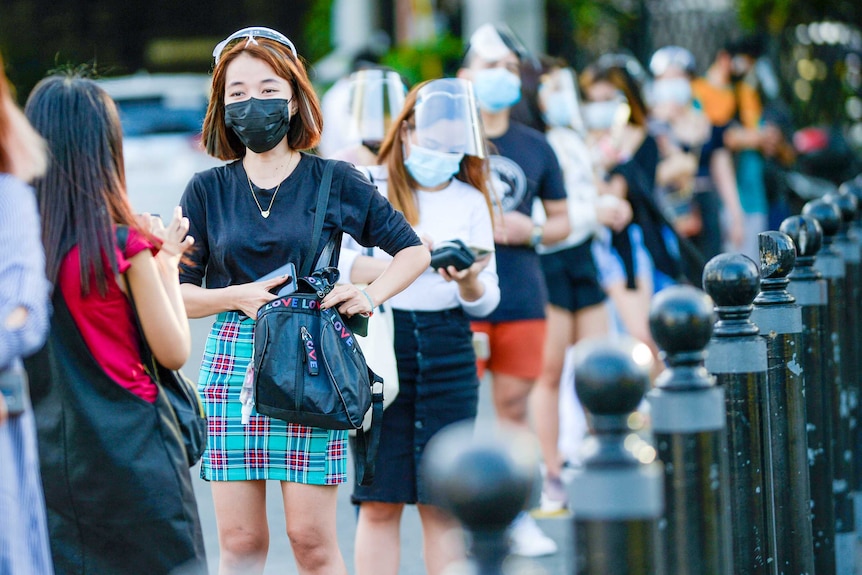 A young woman in a black face mask talk to someone while they line up with other masked people