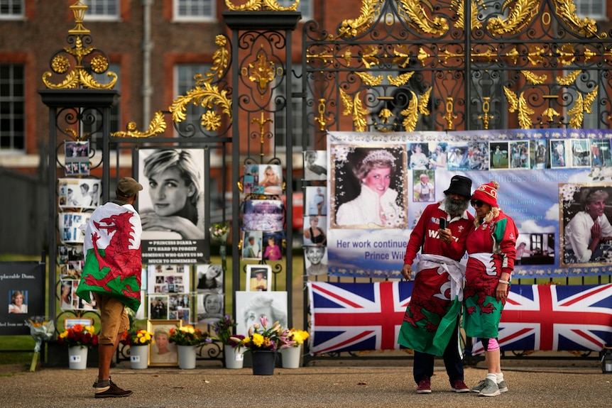 Three people in Welsh flags stand outside the gates where there is a tribute to Princess Diana. 