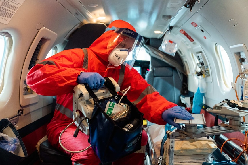 An RFDS doctor decked out in PPE sits on a plane holding medical equipment.
