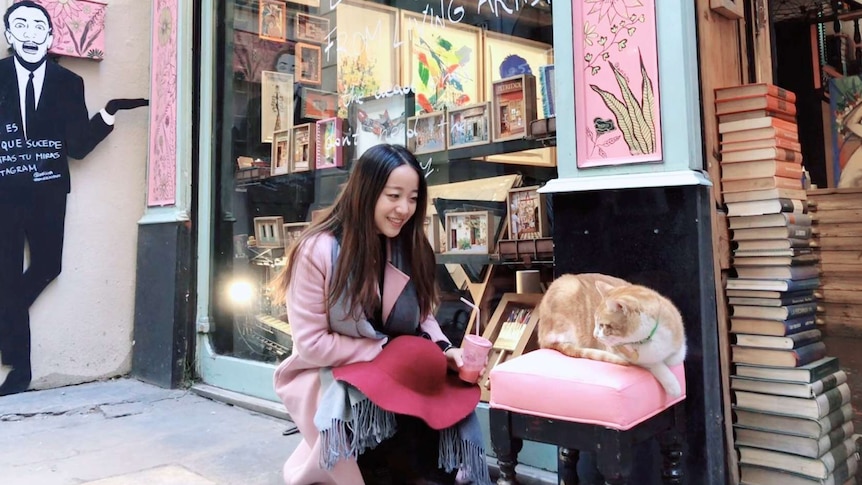 Tong Gong kneels to look at a cat sitting on a chair out the front of a shop, she is smiling.