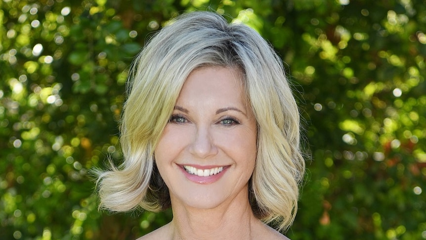 Olivia Newton-John smiles at the camera. She is in front of a blurred green bush