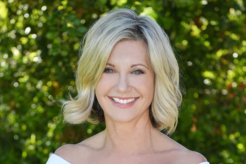 Olivia Newton-John smiles at the camera. She is in front of a blurred green bush