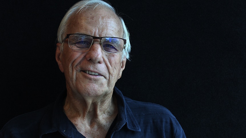 Shot of a white-haired, elderly man standing in front of a black background.