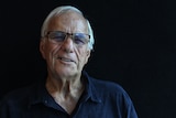 Shot of a white-haired, elderly man standing in front of a black background.