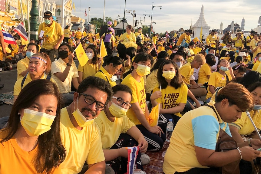 A crowd of people wearing masks and holding yellow flags while wearing yellow shirts sit on the ground in a street.
