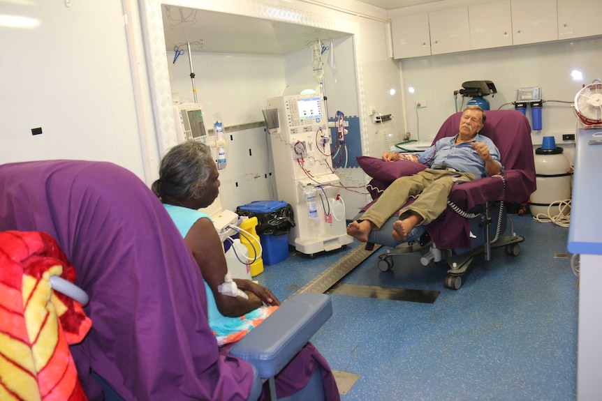 A man sitting in a large chair hooked up to a dialysis machine, chats to a woman