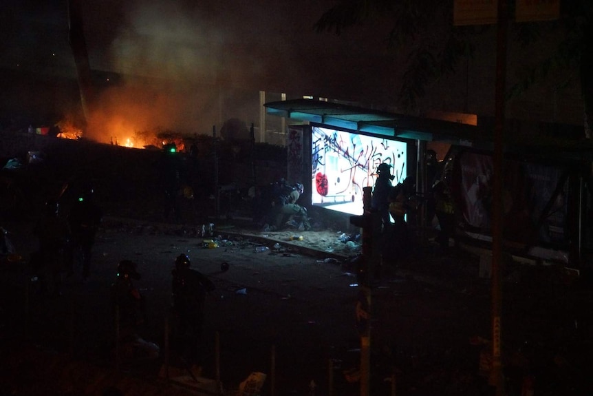 A dark scene with protesters and fire in the background and a lit up sign.