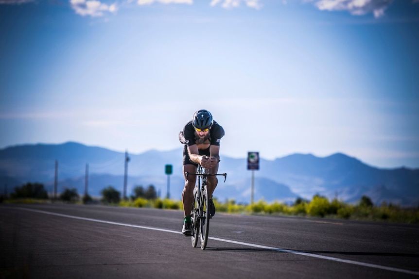 A cyclist rides down an empty road with mountains in the background 