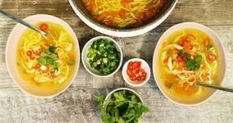 Two bowls of chicken noodle soup on a table with bowls of chilli, spring onion and corianders illustrating our simple recipe