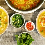 Two bowls of chicken noodle soup on a table with bowls of chilli, spring onion and corianders illustrating our simple recipe