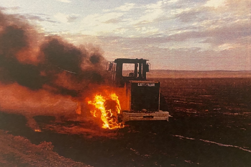 A machine loader vehicle on fire in a field. The front wheel is ablaze and a large plume of dark smoke is rising.