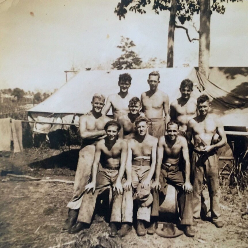 Black and white photo of nine men looking at the camera, shirts off. Tent and bushland in background.