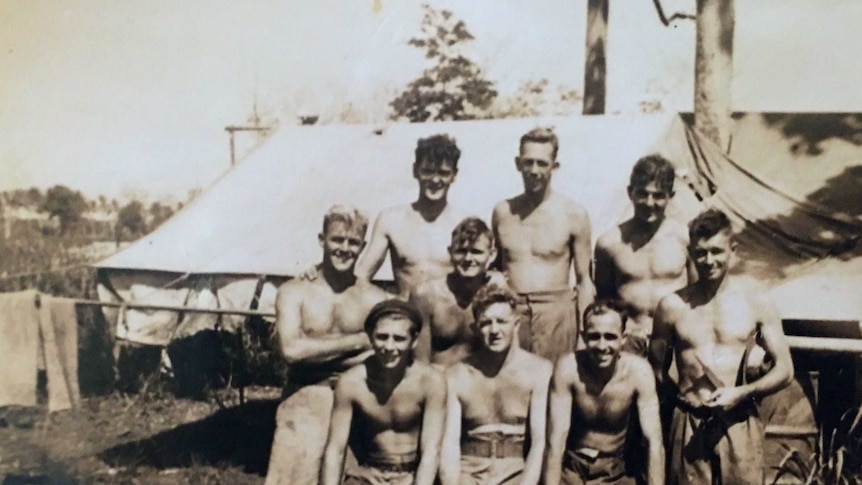 Black and white photo of nine men looking at the camera, shirts off. Tent and bushland in background.