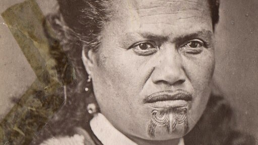 A sepia-toned archival image of a Maori woman with a tattooed chin and upper lip, and feathers in her hair.