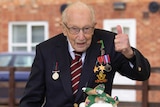 Captain Tom wears glasses, blazer and war medals with a thumbs up in front of cakes decorated with planes and tanks.