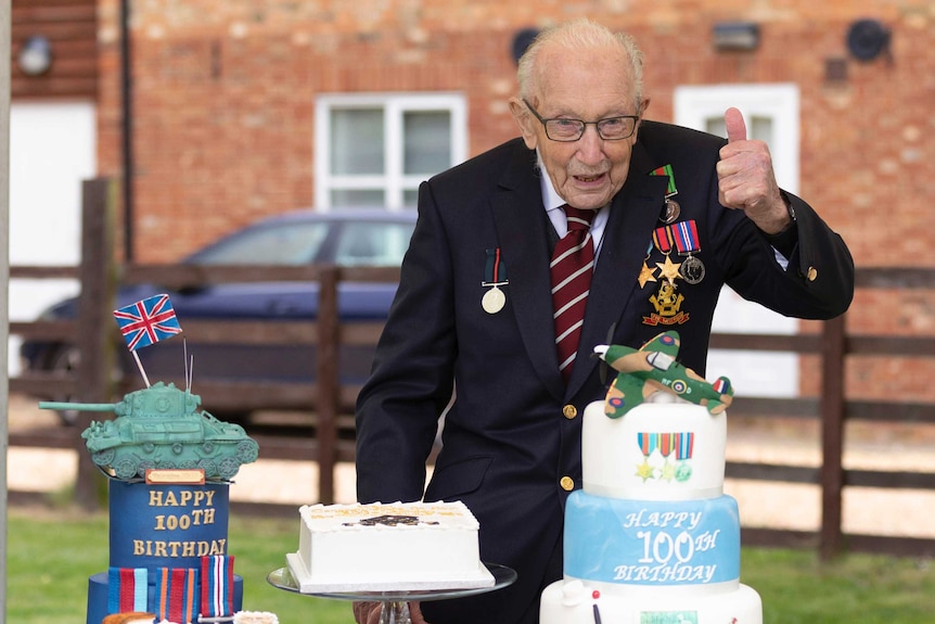 Captain Tom wears glasses, blazer and war medals with a thumbs up in front of cakes decorated with planes and tanks.