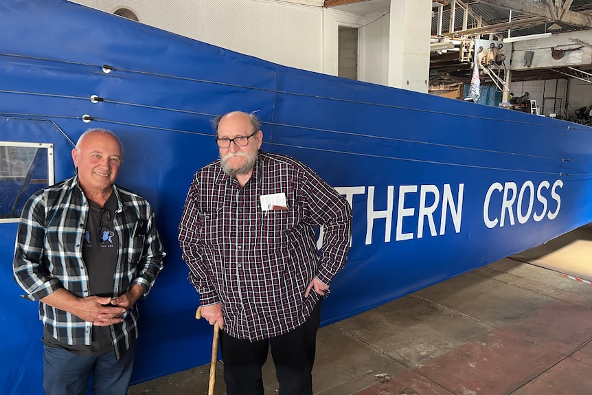Two men stand in front of a replica plane, the words (SOU)THERN CROSS printed on the side of the blue canvas