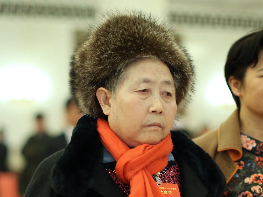 An older woman wearing a bright orange scarf and fur hat frowns, looking slightly unimpressed