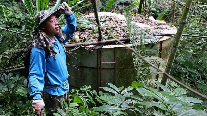 Farmer in long blue shirt and hat rests near a water tank at his tea farm.