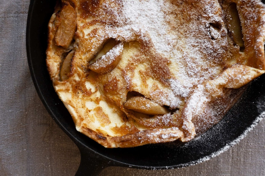 A close up of a just baked puffed pancake with chunks of apple and icing sugar, a simple but special weekend breakfast.