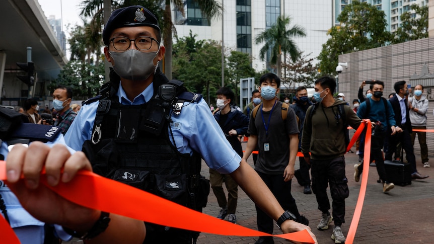 a police officer looking at the camera holds out a red ribbon outside the Kowloon Magistrates Court