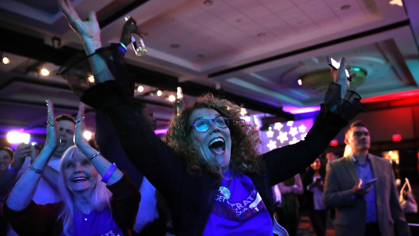 A woman in a blue shirt marked Democrats throws her arms in the air and shouts in celebration at a party