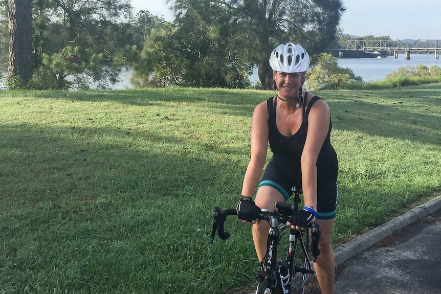 A woman in singlet, cycle pants and on a bike in front of a park with a river in the background