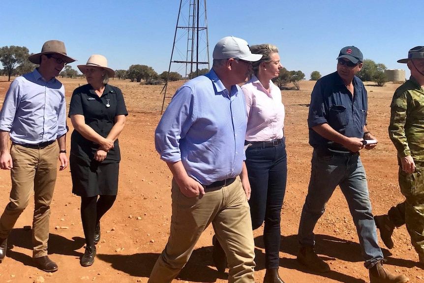 Scott Morrison in a cap, shirt with sleeves rolled up, chinos and boots, walks among a crowd on red dirt in front of a windmill.