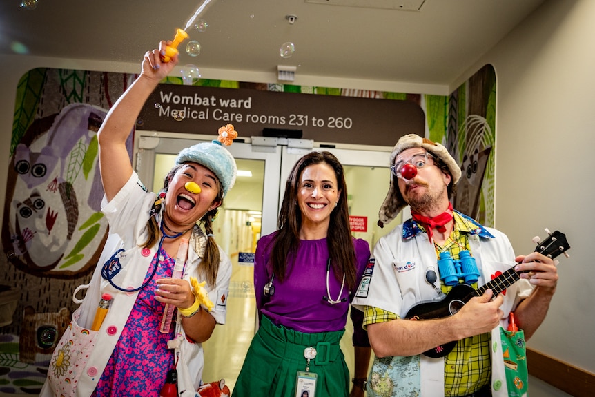 A white woman with long dark hair in a hospital. She's smiling and standing next to two clowns