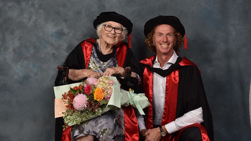 90 year-old woman in graduation cap and gown holds a flower bouquet and smiles next to a man